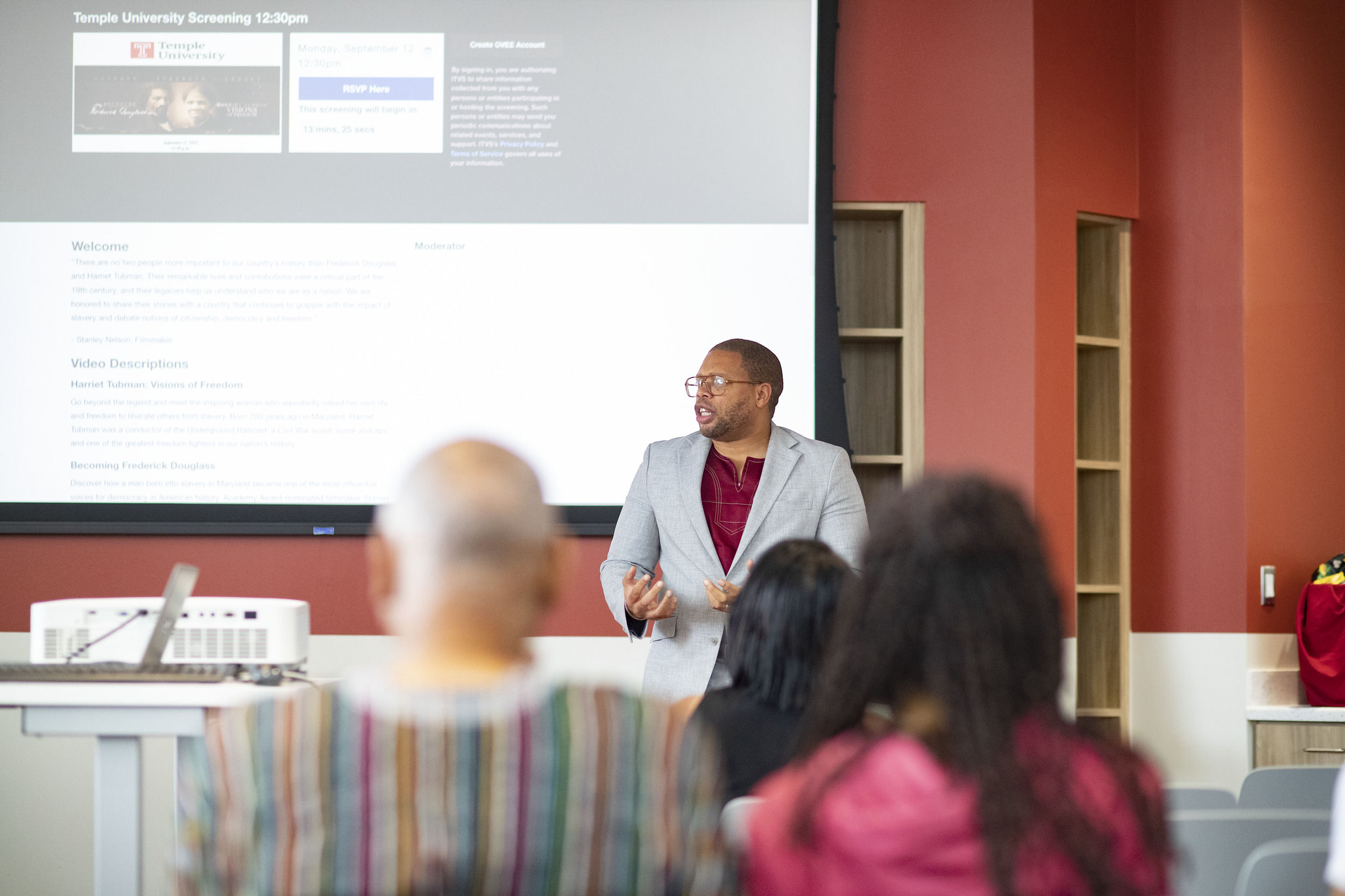 A man at the front of a room, behind a projector, speaks to a group of seated people.