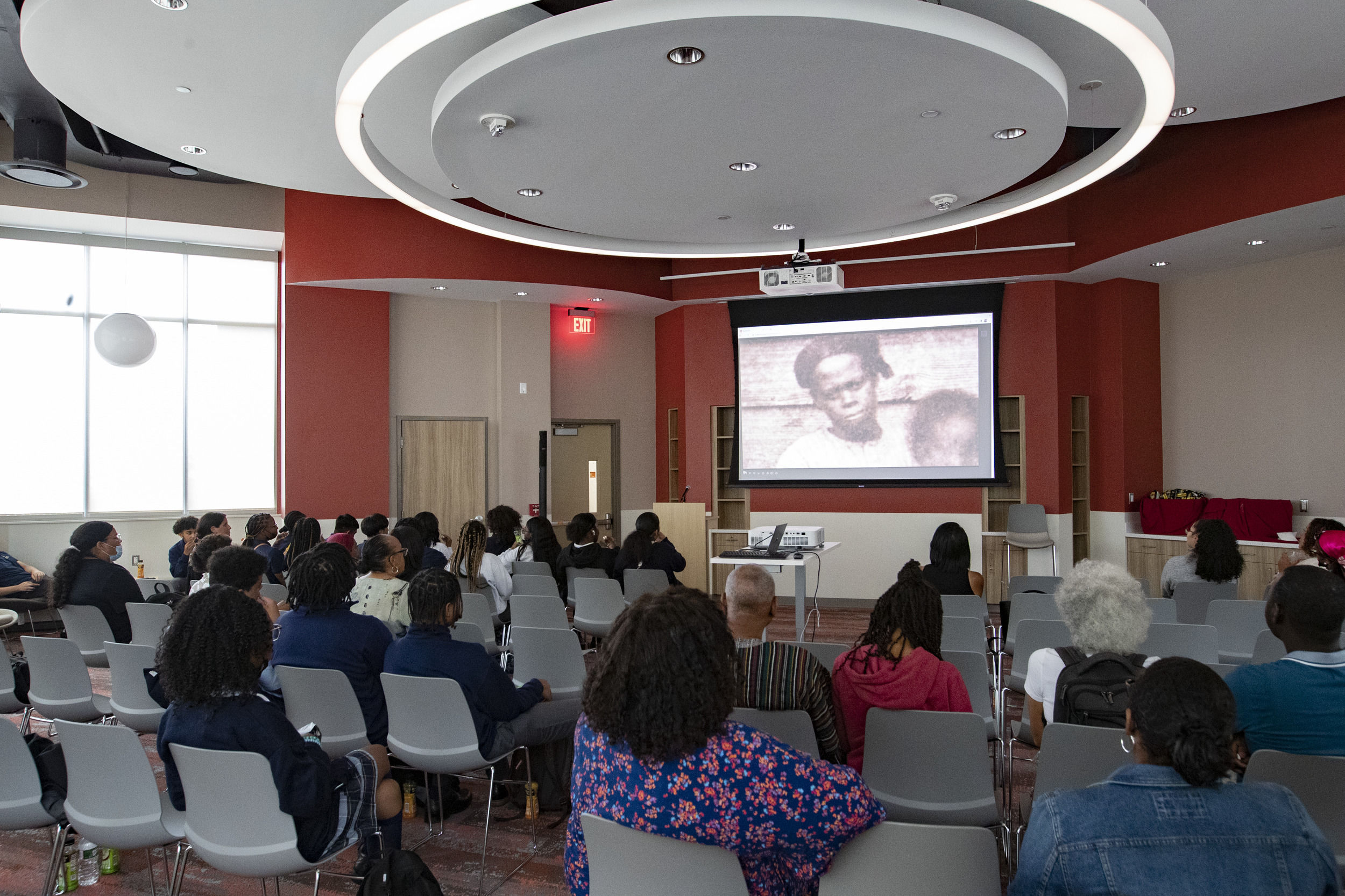 People sit in chars and watch a screen at the front of the room.