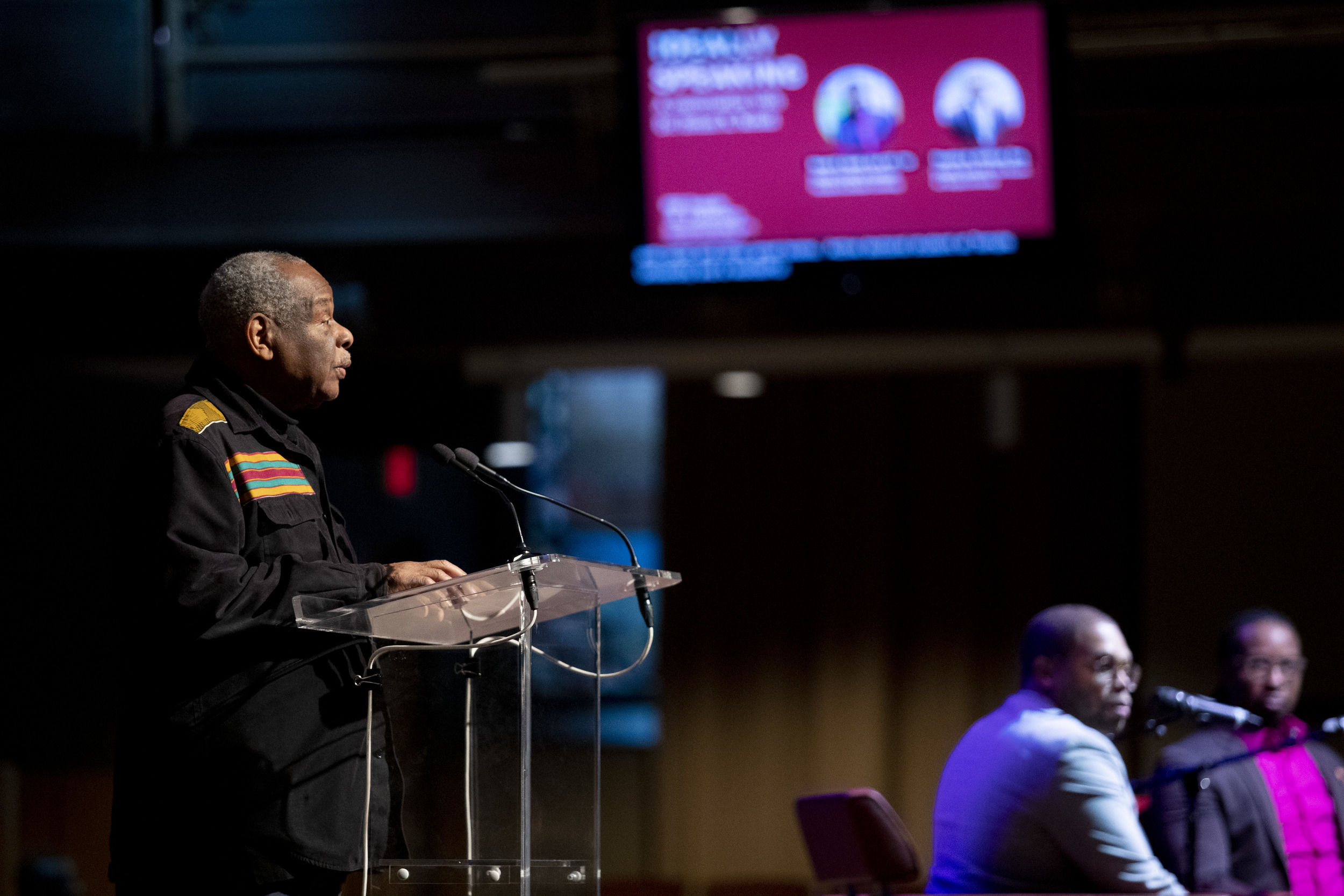 A speaker at a podium with two men sitting on chairs in the background.