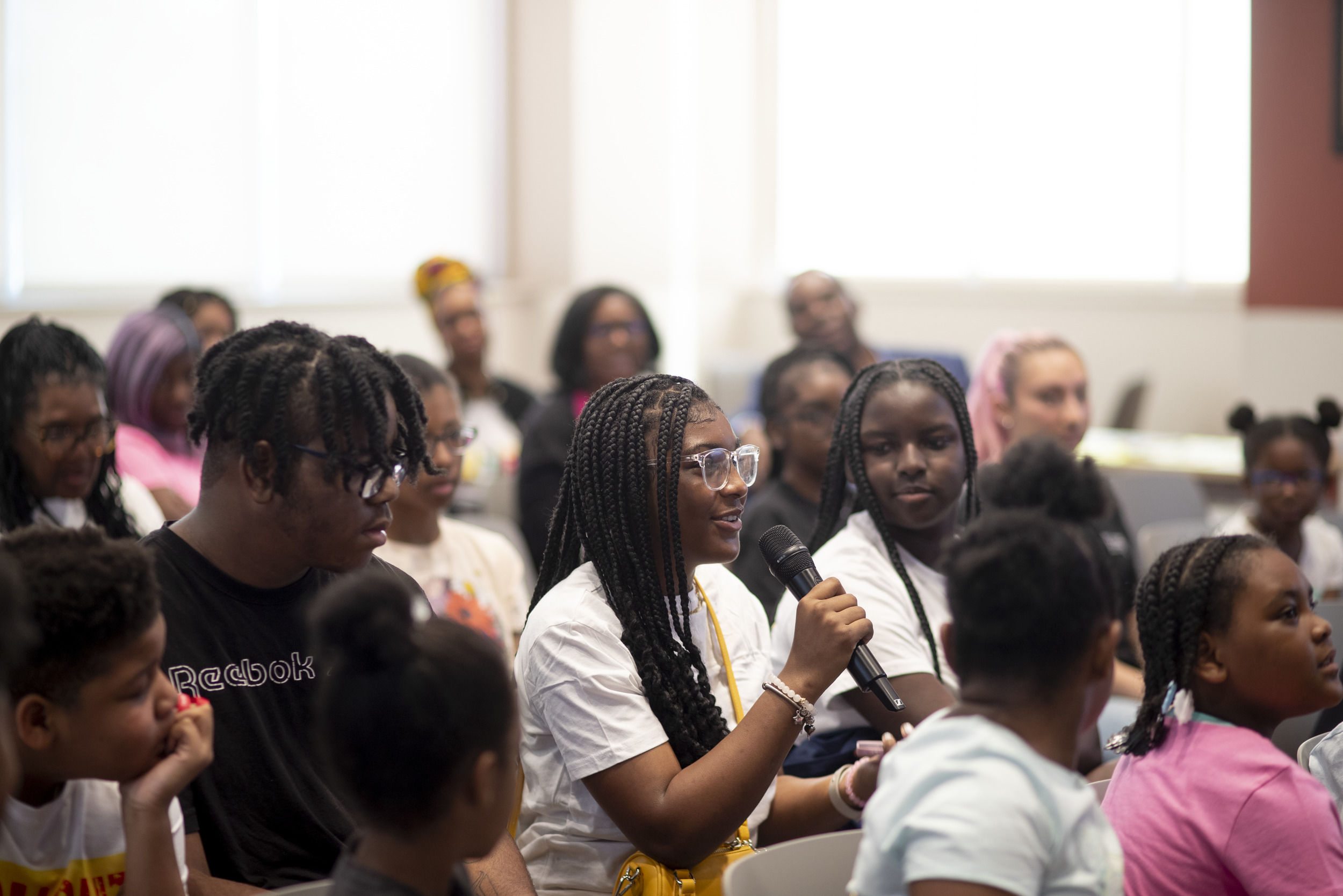 A young girl sitting amongst her peers is speaking into a microphone.