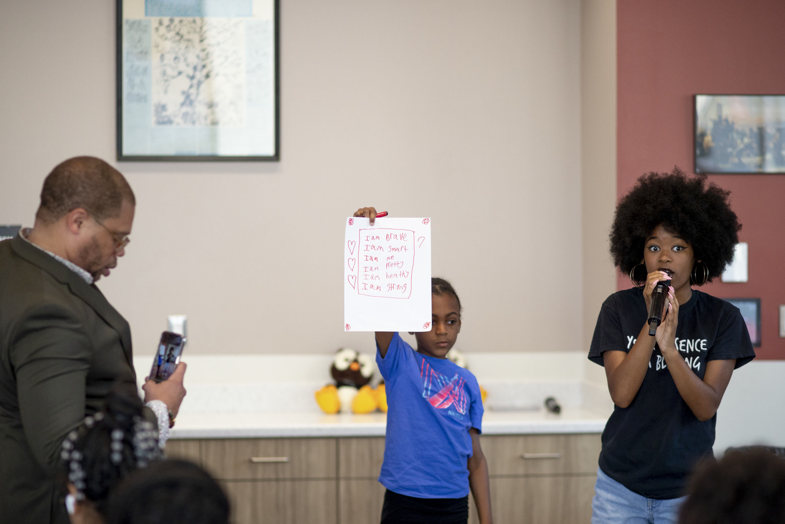 A young child stands in the front of a room while holding up a white piece of paper with writing on it.