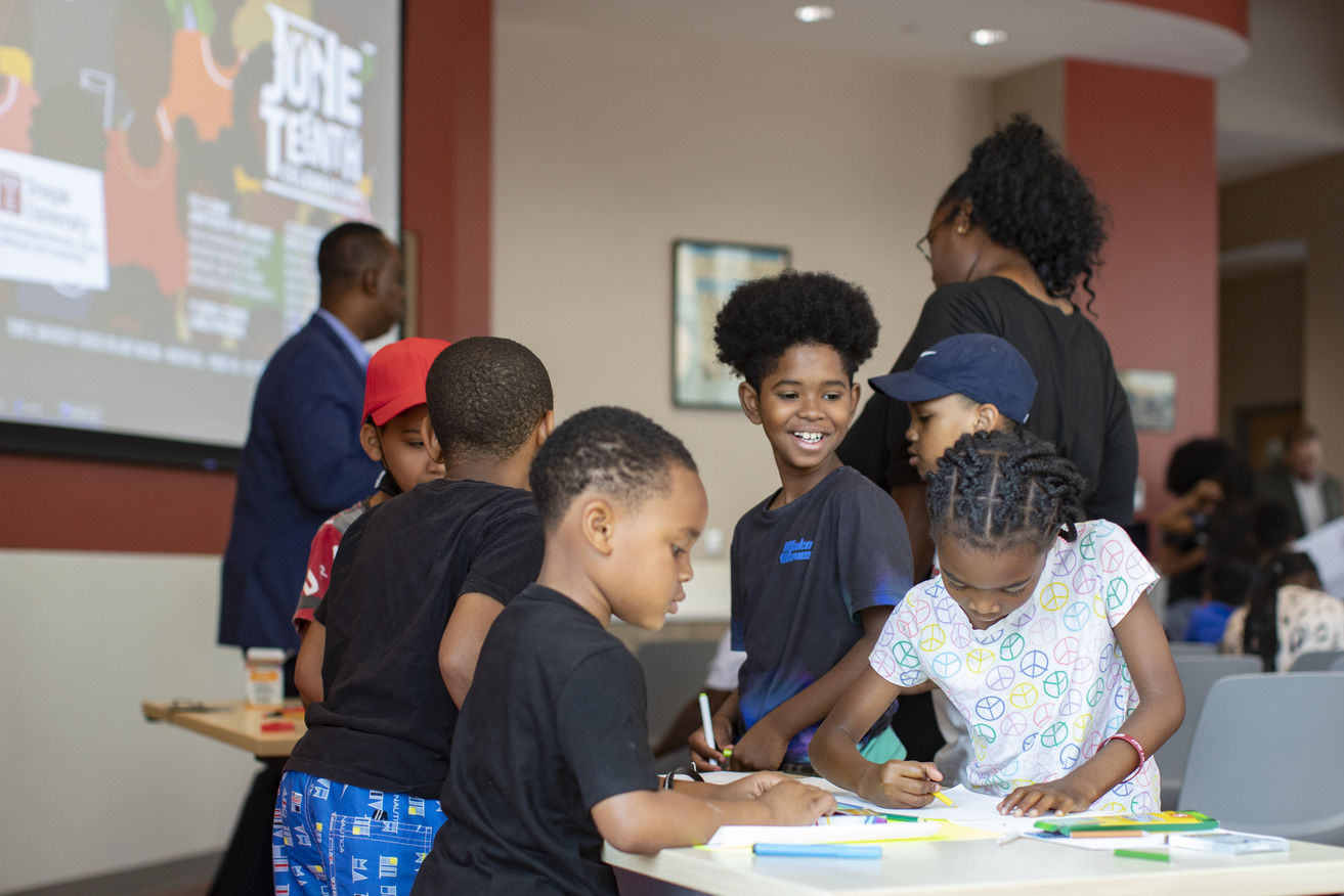 Children stand around a table while coloring and writing on paper.