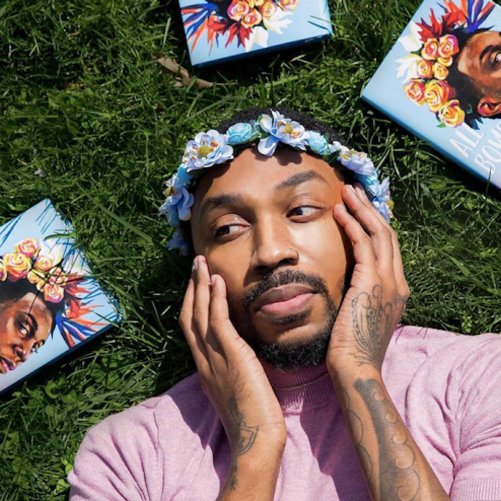 author George M. Johnson wearing a pink t shirt and flower crown, surrounded by their books. They have brown skin with dark hair.