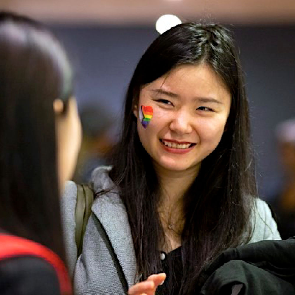 Two temple Students at an Institutional Diversity LGBTQIA + event. One is smiling and has a heart sticker on her right cheek of the LGBTQIA + flag. The student has long straight black hair and light skin
